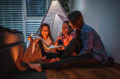 1266844774 A mom with her two daughters camp inside their home due to the coronavirus restrictions and quarantine.They have pitched a tent along and have a fake campfire next to their tent. They are preparing marshmallows on campfire. They are making the best of ...