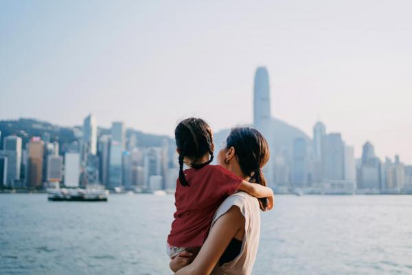 1331476218 Loving young Asian mother embracing adorable little daughter in arms while looking over the city by the promenade of Victoria harbour. Enjoying the spectacular urban city skyline at sunset. Sharing intimate bonding time together