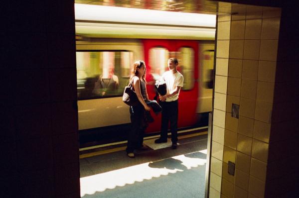 Young male and female standing on platform waiting to board train with red door. 