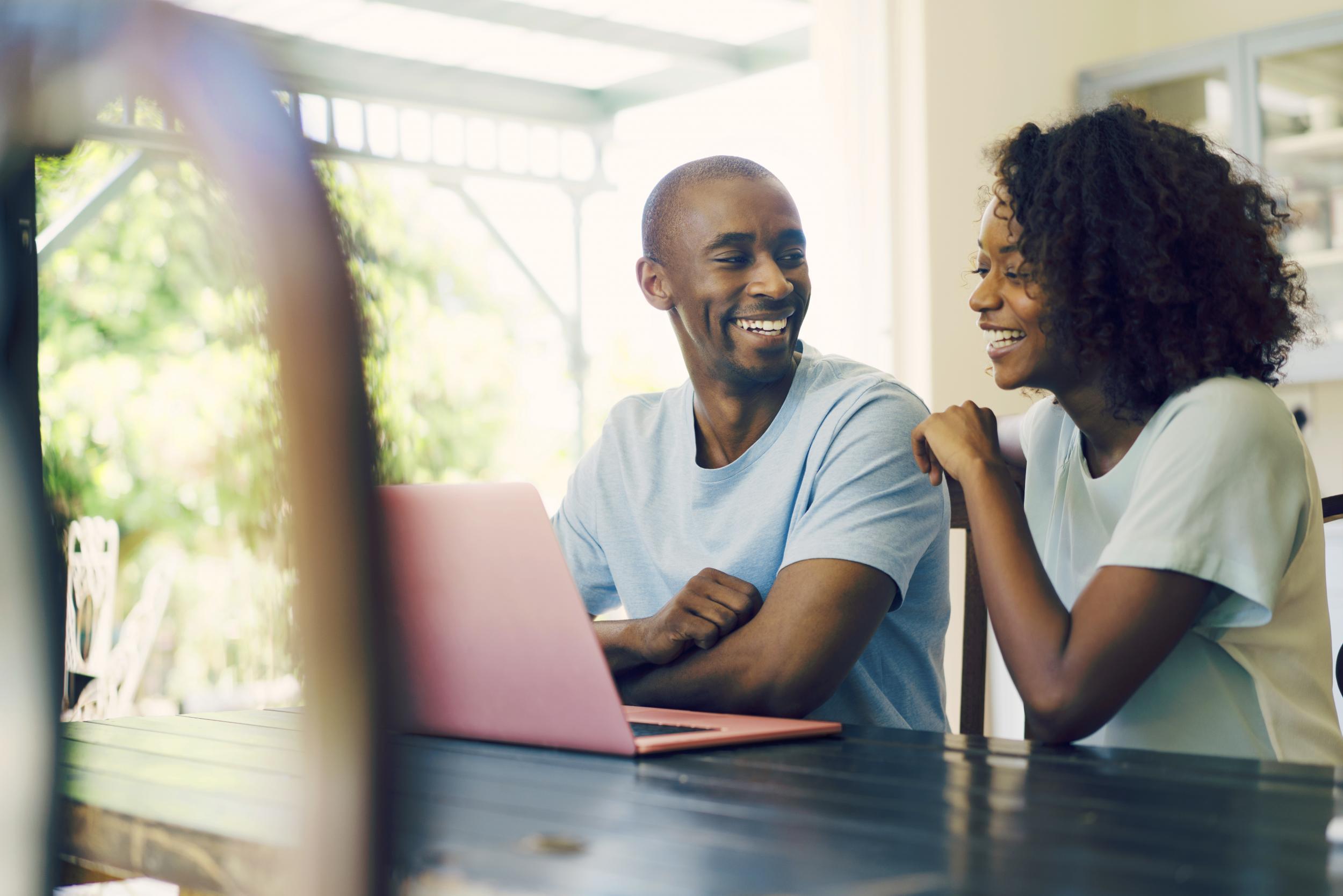 A photo of happy couple using laptop at home. Cheerful man sitting with woman at table. Both are in casuals.