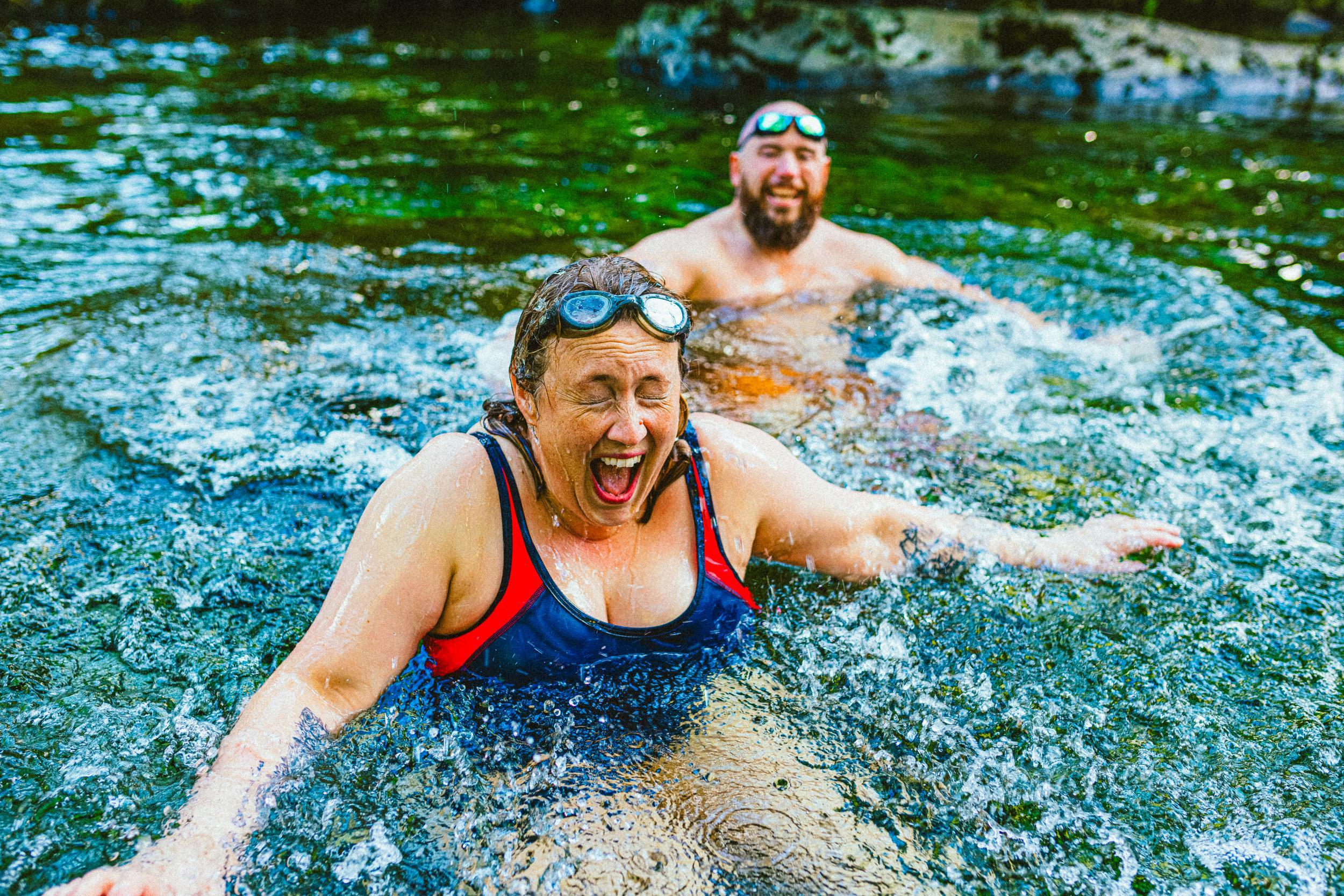 Couple wild swimming in the Lake District, North West of England. They are in a river, enjoying time outdoors. The woman is laughing with her eyes closed and mouth open.