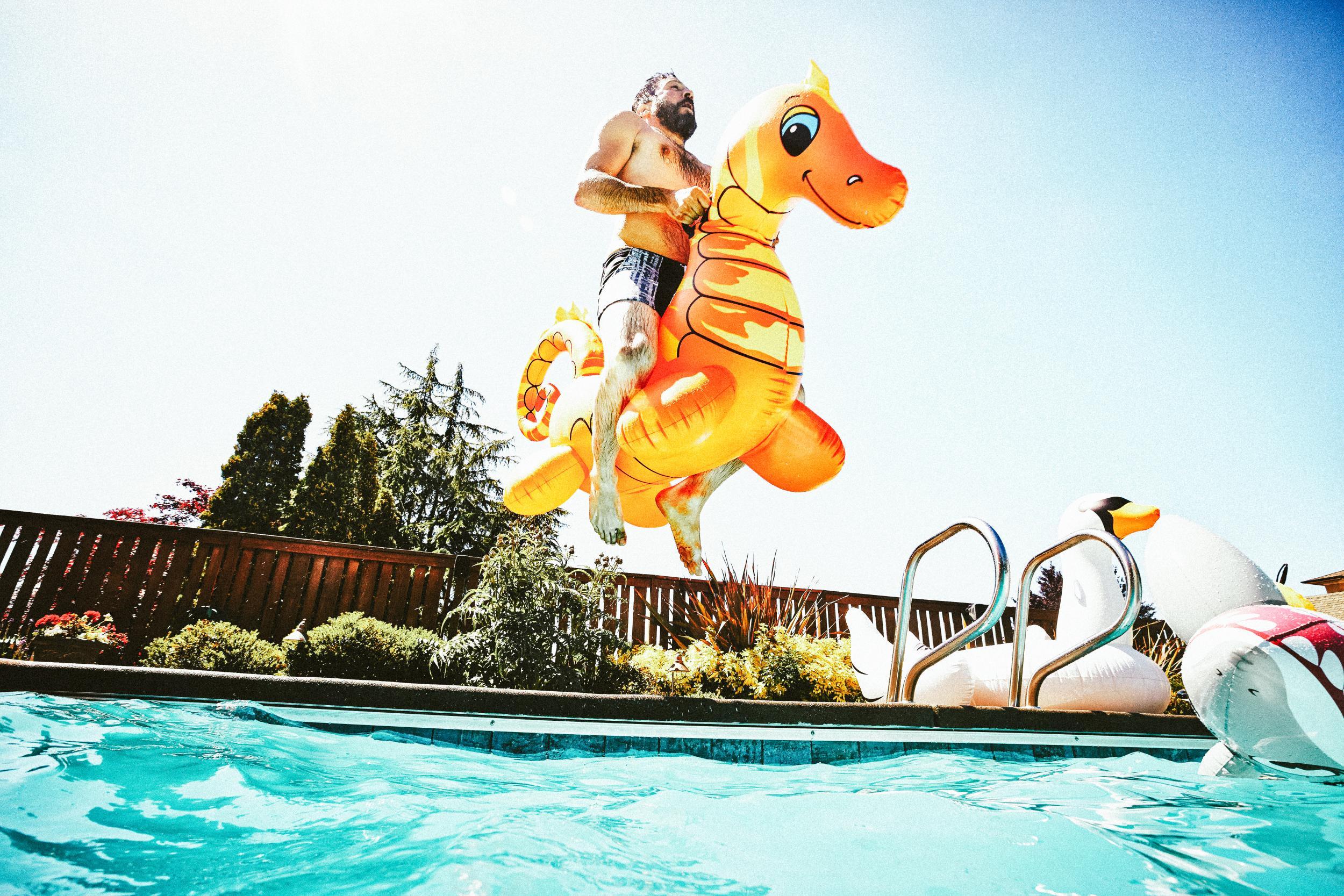 520749695 Man jumping into outdoor pool with inflatable pool toy on summer afternoon