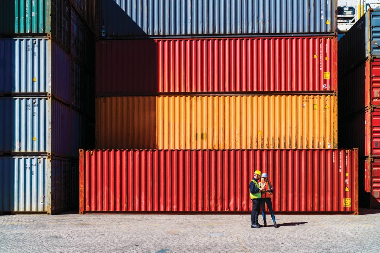 A photo of dock workers having discussion. Full length of manual workers standing against cargo containers. They are working in shipping yard.